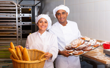 Portrait of successful Hispanic baker couple standing with freshly baked goods in bakery..