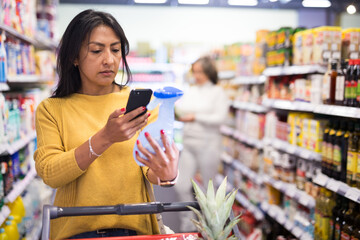 Hispanic woman scanning barcode on detergent bottle with her smartphone while shopping in...