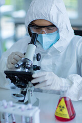 Closeup shot of Asian female professional scientist in ppe full protection suit safety goggles face mask rubber gloves using microscope looking at coronavirus sample with male colleague assistant