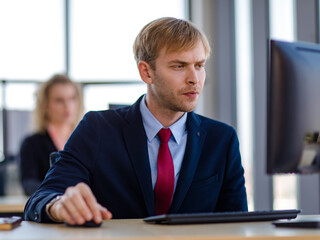 Caucasian stressed depressed upset unhappy blonde hair male businessman employee in blue formal suit with necktie holding looking at smartphone in hand at working desk with computer in company office