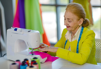 Caucasian old senior professional wrinkled face female designer tailor dressmaker with measure tape use sewing machine stitching colorful fabric textile clothing on table in studio business workshop