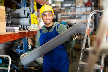 Cheerful Latino workman preparing for pipework routing, choosing supplies in shop of building...