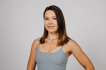 Obraz premium Portrait of a smiling brunette. Beautiful happy young woman in gray top for yoga or fitness poses on gray isolated background in studio, she is standing sideways.