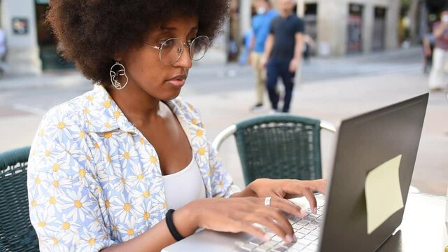 Real time of crop curly haired African American female remote worker in eyeglasses typing on netbook while working in street cafe