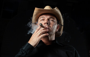 Strong pose of authoritarian man smoking cigar and wearing cowboy straw hat in black shirt, low...