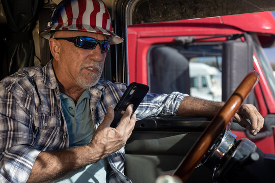 Senior professional truck driver wearing hard hat and sunglasses sits in the cabin and talks on the smartphone. Elderly trucker inside big rig at the steering wheel talking to dispatcher on cell phone