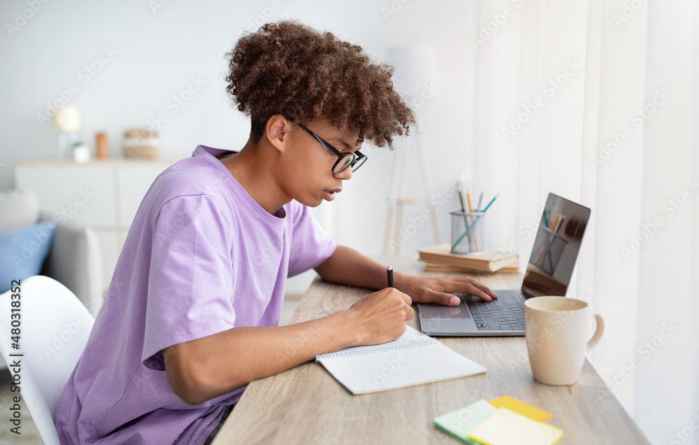 Poster focused black teenager in glasses studying online from home, writing in notebook, using laptop pc at