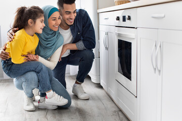 Middle-eastern family sitting near oven in kitchen and looking inside through glass