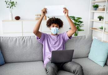 African American youth in face mask showing empty sign with mockup, sitting on couch with laptop during covid