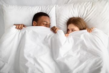 Top view of loving interracial couple hiding under white blanket in bed, looking at each other with...