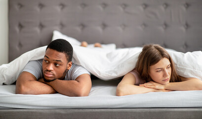 Sad Caucasian woman and her black husband lying apart on bed at home, looking in opposite directions - obrazy, fototapety, plakaty