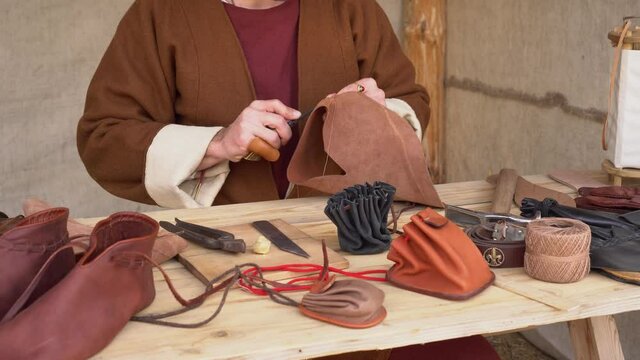 Historical European Leather workshop. Medieval leatherworker craftsmen. Craftsman sews by hand a wallet, shoes, belt, gloves