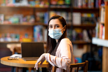 Latin woman in protective face mask looking at camera while sitting in city cafe and working on...