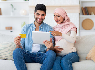 Excited pregnant islamic spouses shopping online with tablet and credit card at home, relaxing together on sofa