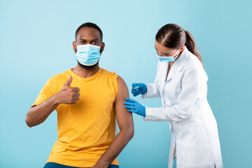 Fototapeta na wymiar Get vaccinated. Young black guy in face mask showing thumbs up during coronavirus vaccination on blue studio background