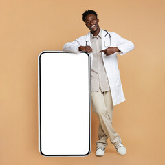Black Young Male Doctor In Uniform Pointing At Smartphone With Blank Screen