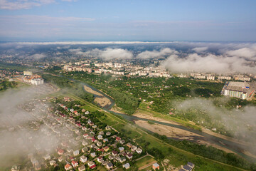 Fototapeta premium Aerial view of white clouds above a town or village with rows of buildings and curvy streets between green fields in summer. Countryside landscape from above.