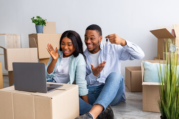 Excited black couple chatting online with friends and showing key of their house to camera, sitting among carton boxes