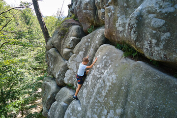 Young child boy climbing steep wall of rocky mountain. Kid climber overcomes challenging route. Engaging in extreme sport concept
