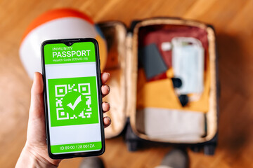 The concept of new normal. Close up view of a male hand holds a smartphone with an immune digital health passport. Top view. A suitcase with clothes and protective masks on the blurred background
