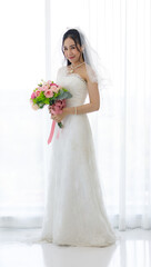 Studio shot of Asian young happy beautiful luxury long hair female bride in white wedding dress standing smiling posing holding pink roses flower bouquet in front of curtain in engagement ceremony