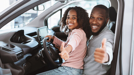 Happy black father and daughter sitting on driver seat of new car, showing thumb up, recommending...