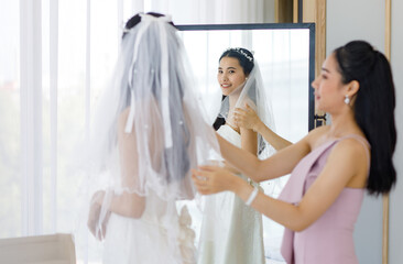 Naklejka premium Asian lovely beautiful happy cheerful female bride in white long wedding dress standing staring at her reflection in mirror while pretty bridesmaid helping adjusting facial veil in blurred foreground