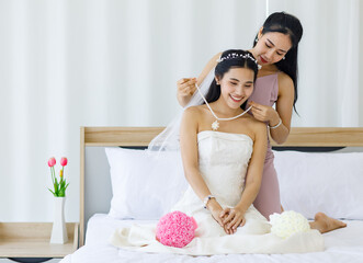 Obraz na płótnie Canvas Asian female bridesmaid helping adjusting dressing hairstyle and facial veil for young beautiful happy bride in white wedding dress sit smiling on bed in bedroom preparing for traditional ceremony
