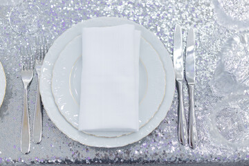 Festive table setting with plates, silverware and white napkin on glitter tablecloth decorations in silver color. Top view