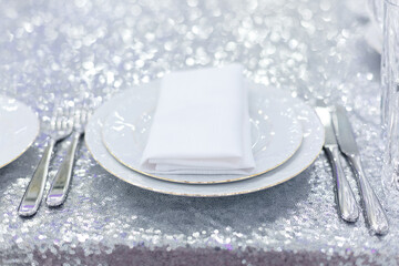 Festive table setting with plates, silverware and white napkin on glitter tablecloth decorations in silver color. Soft focus bokeh