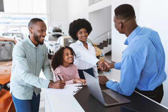Its a deal. Black family buying new car, shaking hands with salesman, signing auto purchase agreement at dealership