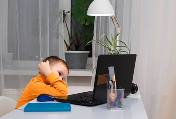 Child is tired of learning. Home schooling, homework. The boy is sad sitting at table with laptop. A bored and sad child looks at the computer, the child is tired of online communication and learning