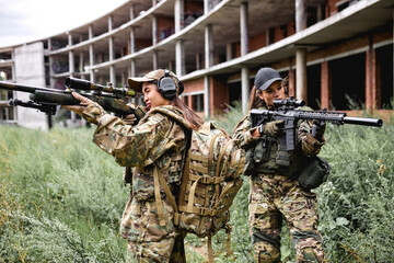 Fototapeta premium Powerful army ladies women in military gear posing for photo in field at summer season. Female is Wearing green camo uniform and weapon with suppressor, abandoned building in the background