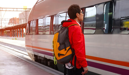 Tourist Asian man in red coat waiting  with backpack in the railway, which Backpack  the train station with a traveler, Travel concept