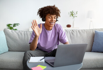 Happy black teenager in headphones greeting his friend or teacher online, waving at camera from home - obrazy, fototapety, plakaty