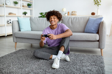 Full length of positive black teen gamer with controller playing online video games, sitting on floor at home © Prostock-studio
