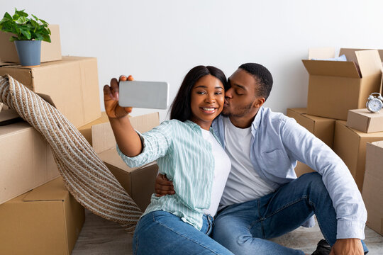 Happy black spouses taking selfie in their new apartment, celebrating moving day, sitting near carboard boxes