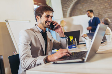 Happy businessman using laptop and phone with team discussing project in the background.