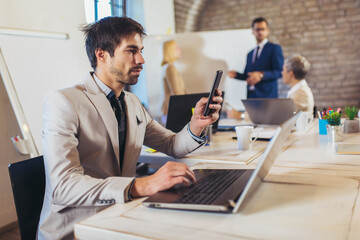 Happy businessman using laptop and phone with team discussing project in the background.