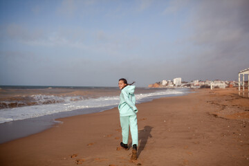 Teenager female walking outdoors in the beach in spring weather with wind, look to the sea, Girl dreased in blue hoody sweater and trousers