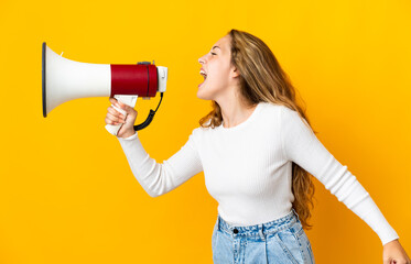Young blonde woman isolated on yellow background shouting through a megaphone to announce something in lateral position