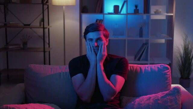 Unbelievable situation. Shocked man. Meme expression. Confused guy grasping face with hands looking amazed sitting sofa neon light room interior looped.