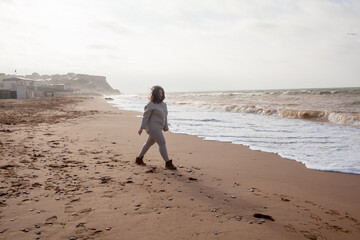 Beautiful woman wearing casual clothes, biege leggings, walking in windy sprin cold weather in the beach against the ocean.  Overweight woman have fun and enjoy the mood of view