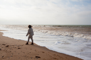 Beautiful woman wearing casual clothes, biege leggings, walking in windy sprin cold weather in the beach against the ocean.  Overweight woman have fun and enjoy the mood of view