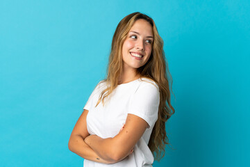 Young blonde woman isolated on blue background with arms crossed and happy