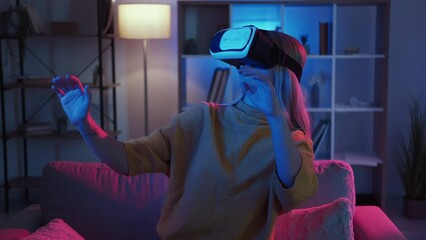 Female gamer. Virtual reality. New experience. Amazed woman in vr goggles showing surprise playing cyber game sitting sofa dark neon light home interior.