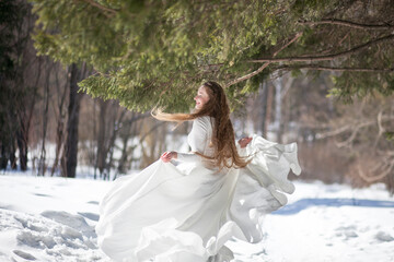 Fototapeta na wymiar blonde with long hair, a girl in a long white dress walks in the winter forest, the snow queen, the bride with a bouquet of dianthus