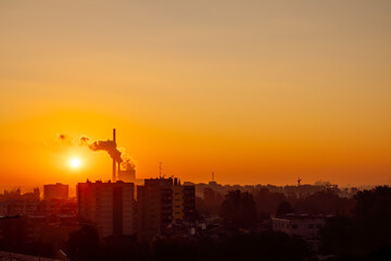 October sunrise over the city of Krakow, Poland, silhouettes of buildings and rising smoke from a thermal power plant, orange colored sky in the background.