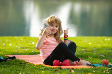Healthy lifestyle and healthy food concept. Little boy child in sportswear with bottle water with thumb up sitting on sport mat after training on sunny spring day. Concept of children sport.