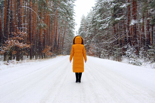 A young woman in a yellow stylish down jacket with a hood stands in the middle of a road in a forest in winter during a snowfall. Beautiful cold season with frost. Holiday weekend in nature.